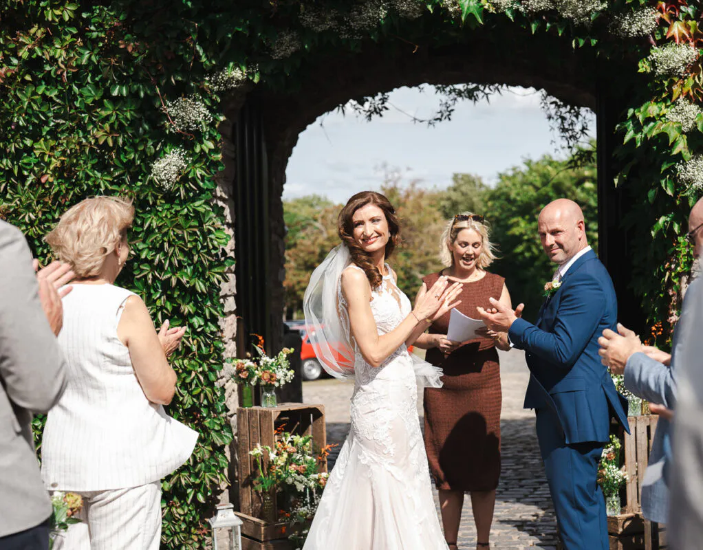 A newlywed couple beams and applauds while walking through a stone archway covered in lush green ivy, surrounded by cheering guests during an outdoor ceremony.