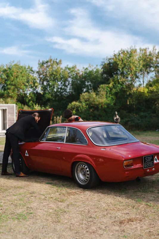 A groom in a black suit looks under the bonnet of a gleaming vintage red classic car parked outdoors on a grassy lawn.