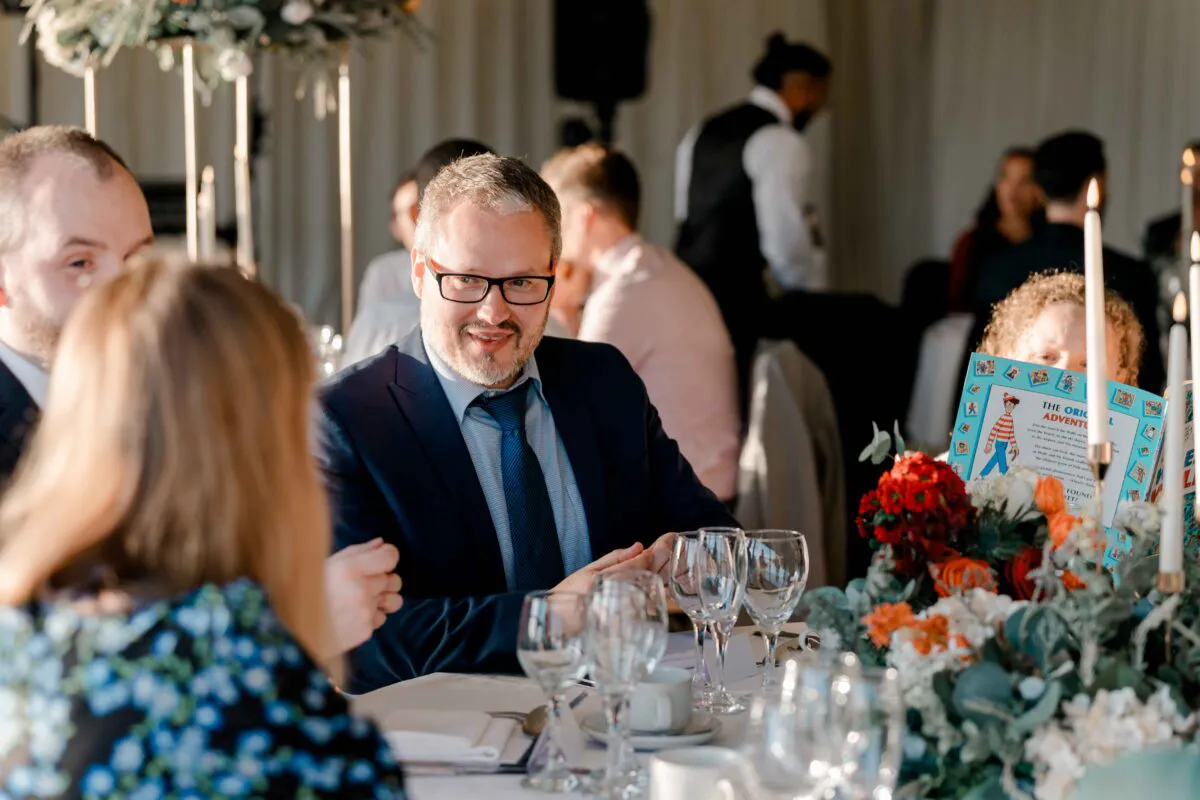 A smiling guest in a navy suit and glasses enjoys a lively wedding breakfast at a table adorned with red floral centrepieces and taper candles inside a marquee.