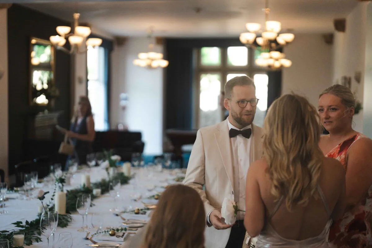 A groom in a beige suit and black bow tie chats with guests near a long banquet table decorated with greenery inside an elegant reception venue.