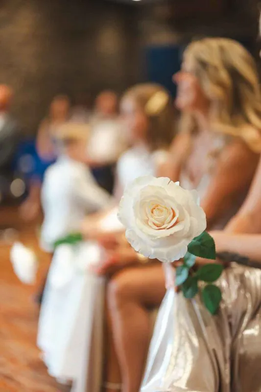 A single white rose is held in the foreground with a shallow depth of field, while wedding guests sit blurred in the background.