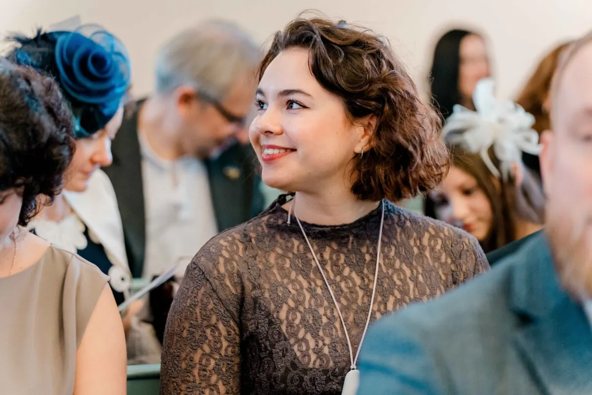 A smiling wedding guest with short curly brown hair and a lace top looks attentively towards the ceremony amidst a seated congregation.