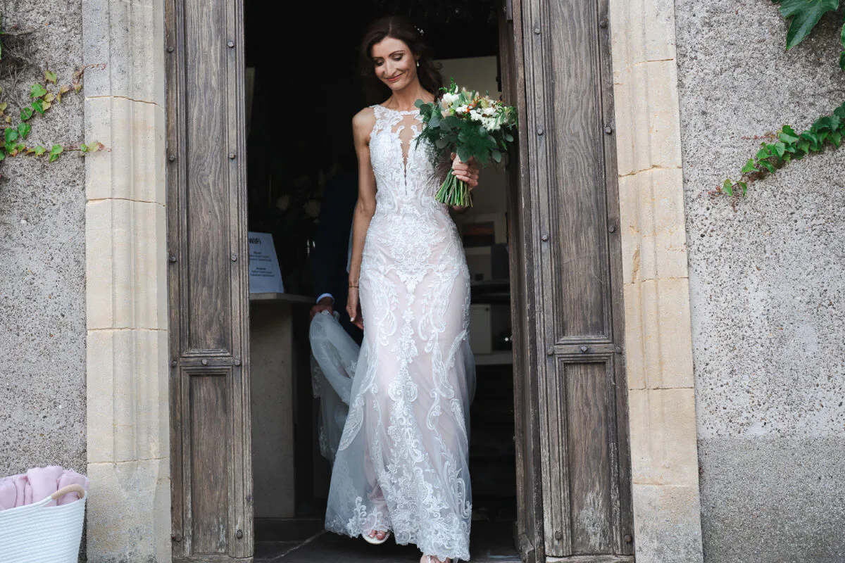 A smiling bride holding a bouquet steps through a rustic wooden doorway framed by stone and ivy.