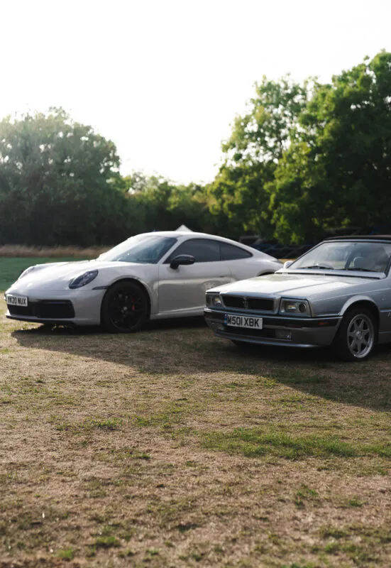 Two luxury sports cars parked side by side on a grassy field under the warm evening sun.