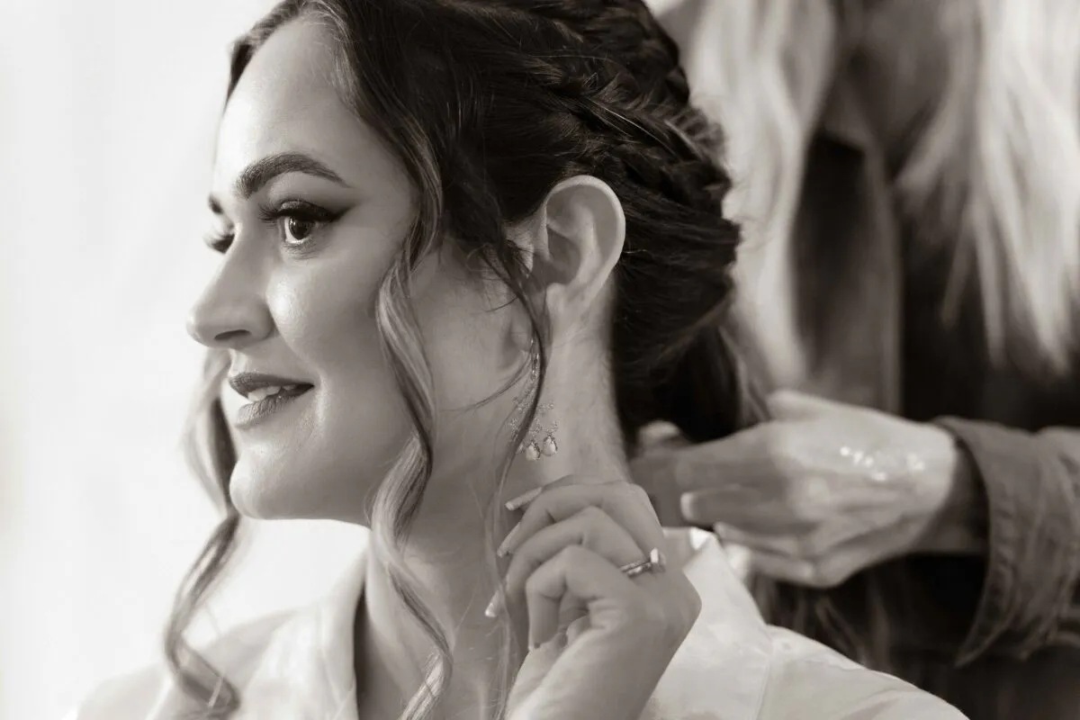 A close-up black and white shot of a smiling bride getting her hair styled during wedding morning preparations while wearing delicate earrings and her engagement ring.
