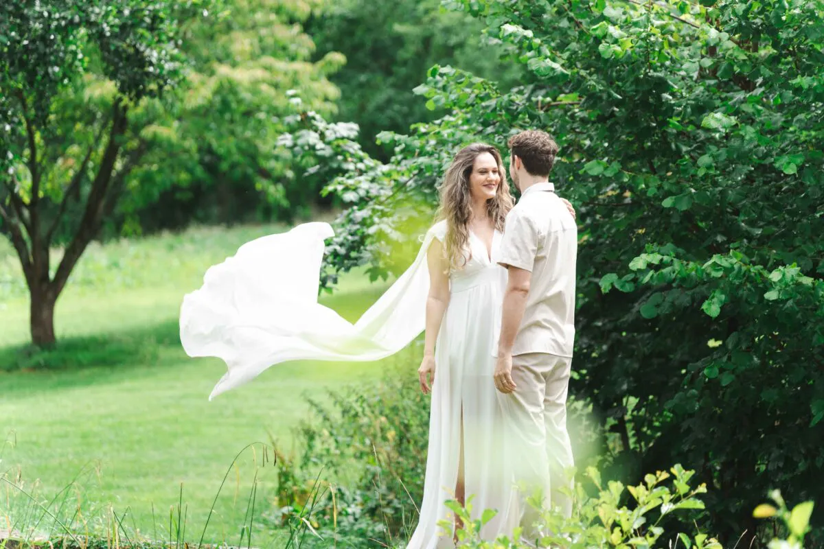 A newly married couple stands together in a lush garden as a light breeze catches the bride's dramatic white tulle cape, creating a flowing motion effect.