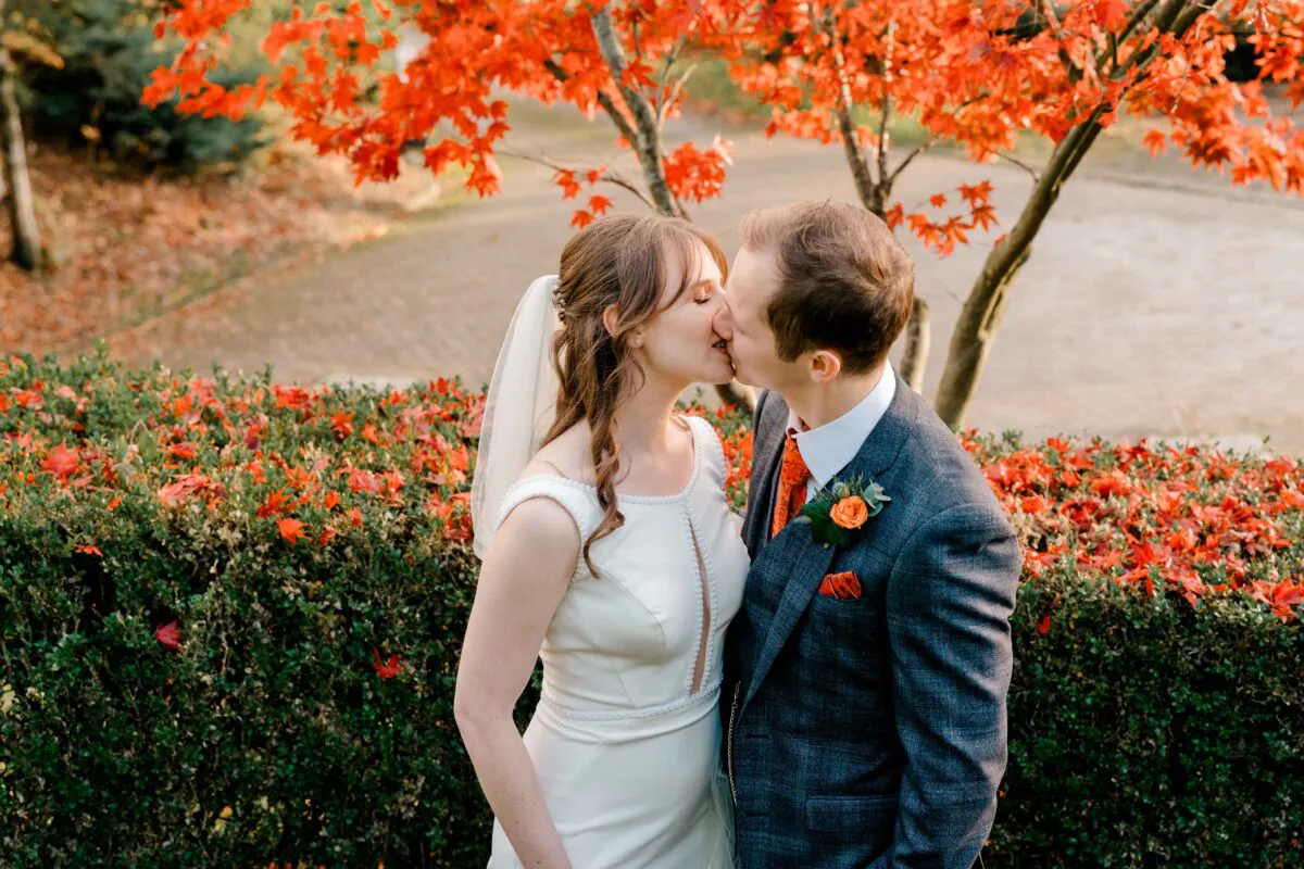 A newlywed couple shares a tender kiss outdoors against a backdrop of vibrant autumn leaves and evergreen hedges during their wedding portrait session.