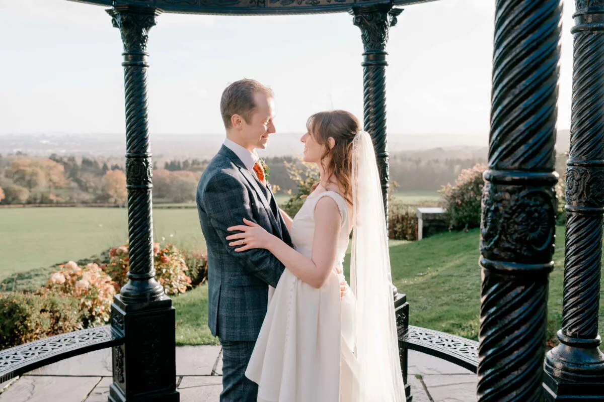 A newlywed couple stands face-to-face embracing inside a dark iron bandstand overlooking a scenic landscape of rolling hills during golden hour.