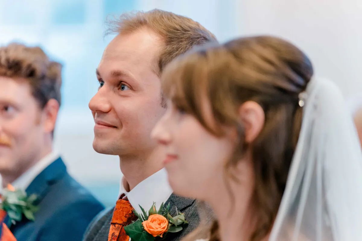 A smiling groom wearing a grey suit and orange patterned tie looks attentively forward alongside his bride during the wedding ceremony.