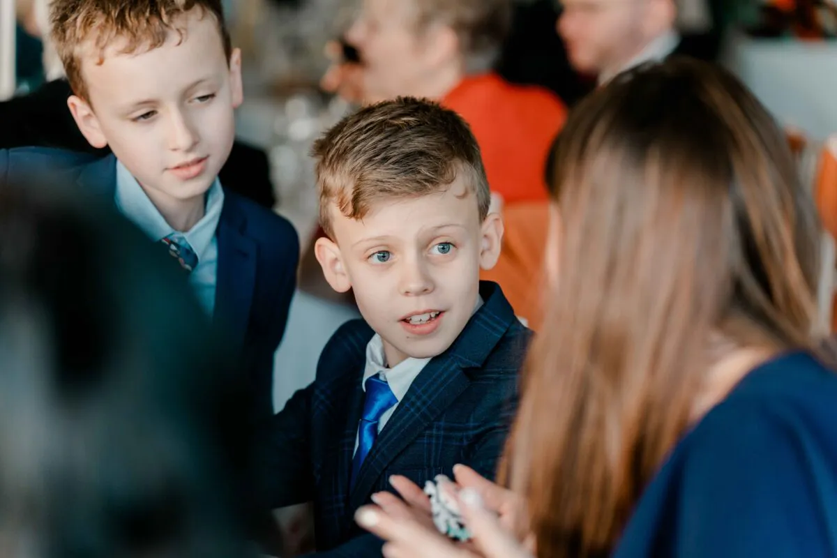 Young pageboys in matching blue suits and ties engage in a lively conversation with a guest during the wedding reception.