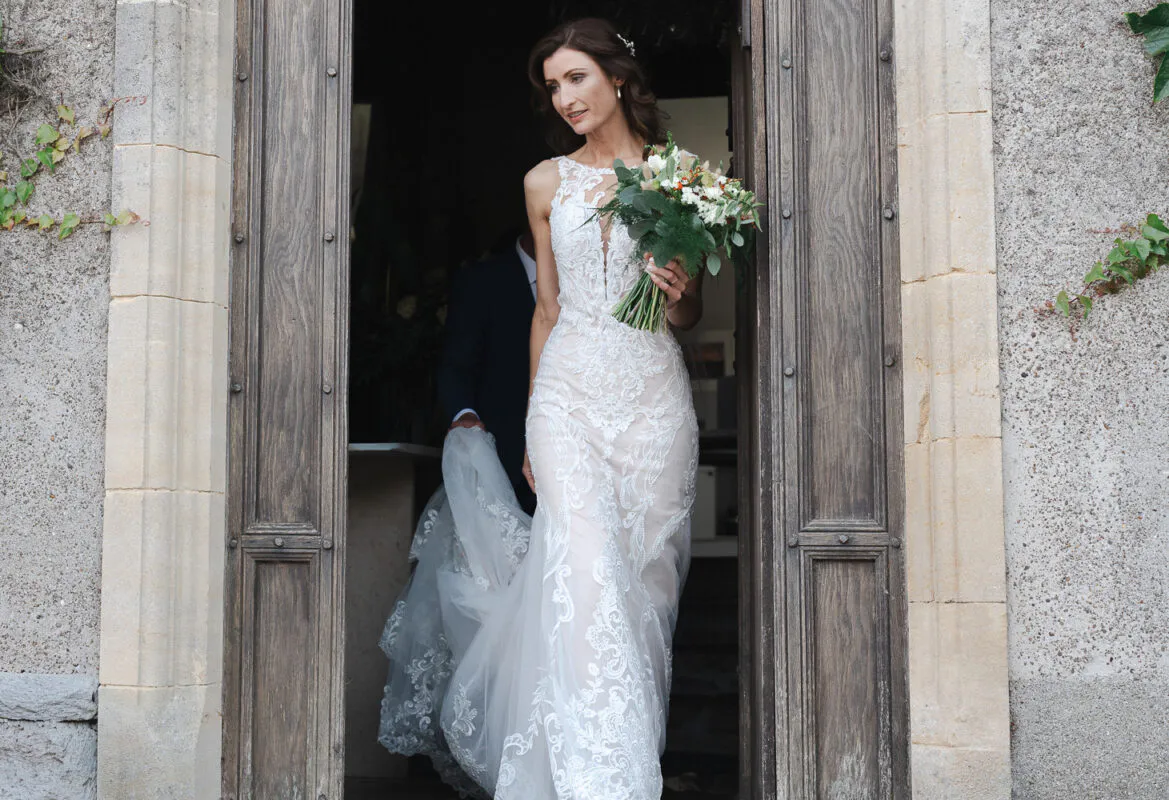 A stunning bride holding a hand tied bouquet steps through a rustic wooden doorway framed by stone masonry on her wedding day.