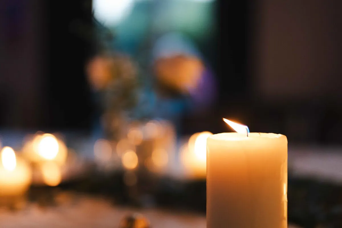 A close-up shot of a single lit pillar candle with a warm, glowing flame against a soft-focus bokeh background filled with candlelight.