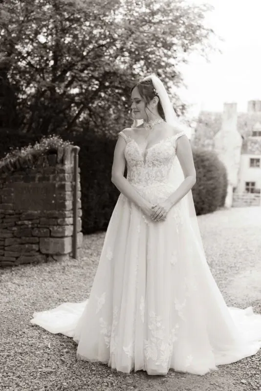 A sepia-toned bridal portrait of a bride wearing an off-the-shoulder lace a-line gown and a veil, standing in a cobbled courtyard outside a historic manor house.