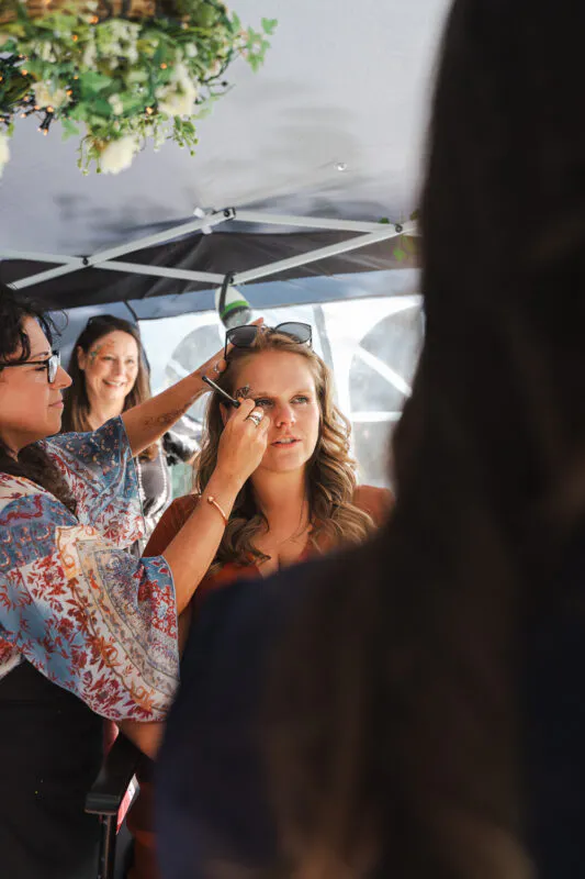 A makeup artist applies detailed face paint or festival makeup to a woman under a canopy during wedding preparations.