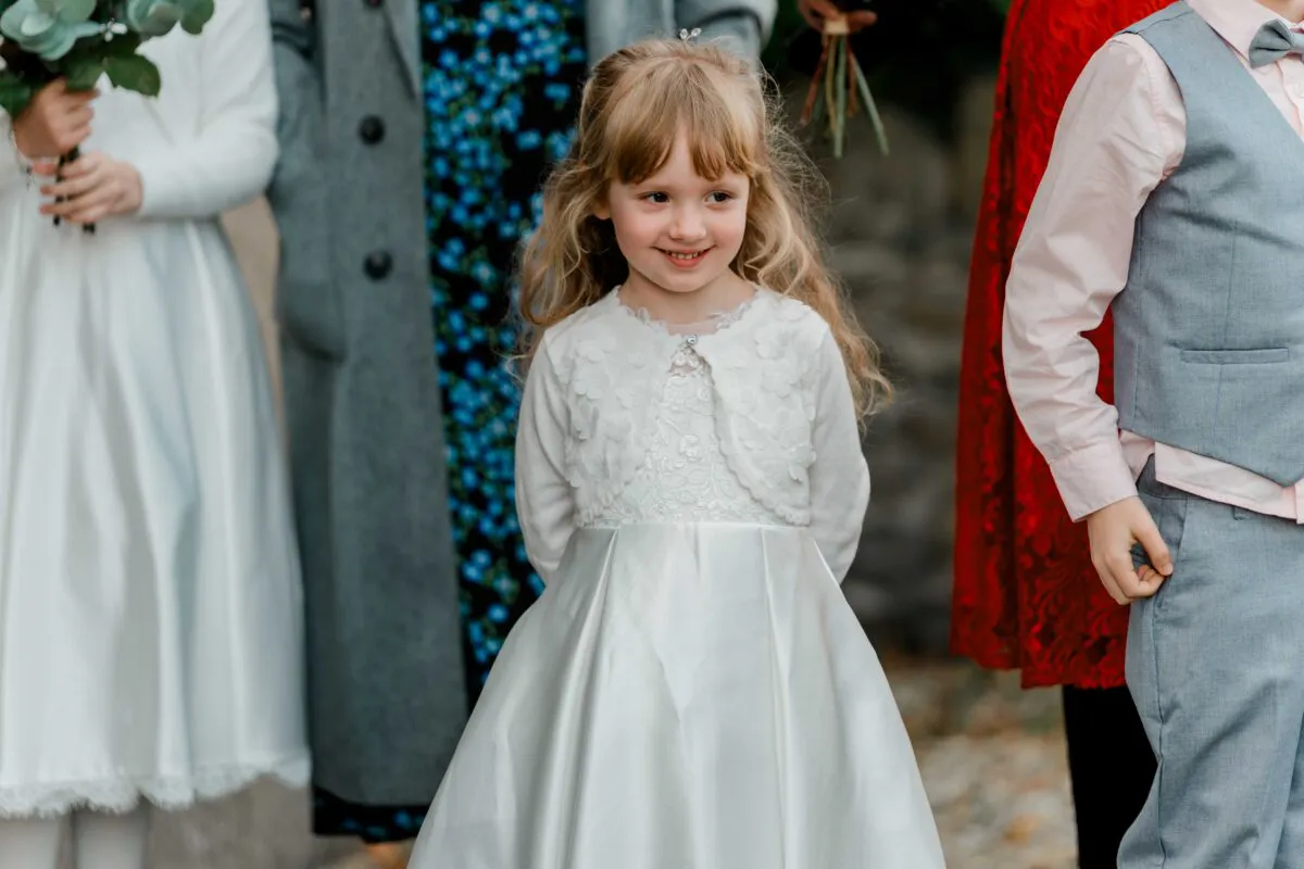 A young flower girl with long blonde hair smiles gently while wearing a white dress and matching cropped cardigan among wedding guests.