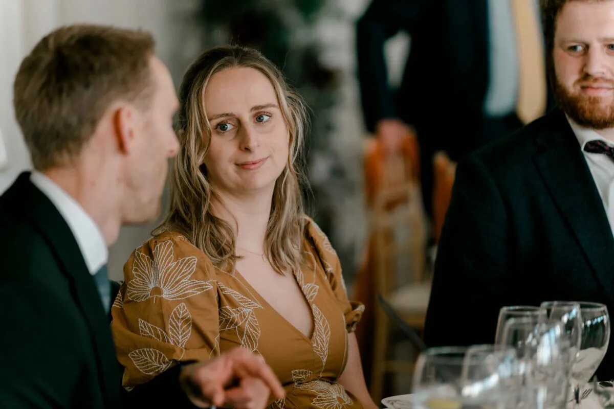 A wedding guest in an ochre patterned dress listens intently during the wedding breakfast while seated at a table with glassware.