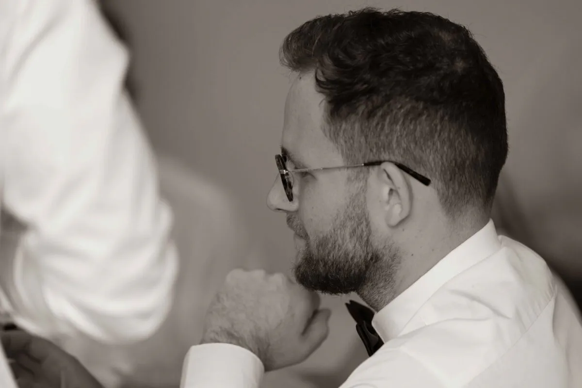 A candid, sepia-toned shot of a bearded groom in a white shirt and bow tie, wearing glasses and looking thoughtfully to the side.