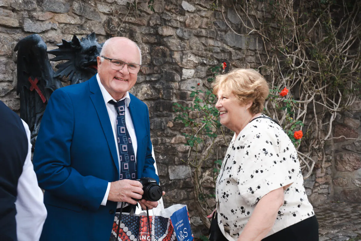 An older man in a blue suit holding a small camera and a woman smiling happily stand together outdoors in front of a stone wall.