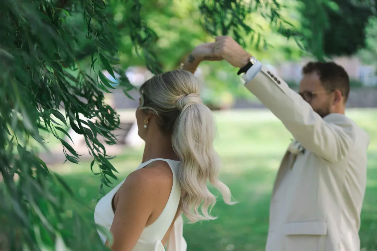 A newlywed couple share a tender dance outdoors near a weeping willow tree, with the bride seen from behind wearing a stylish low ponytail and a modern white dress.