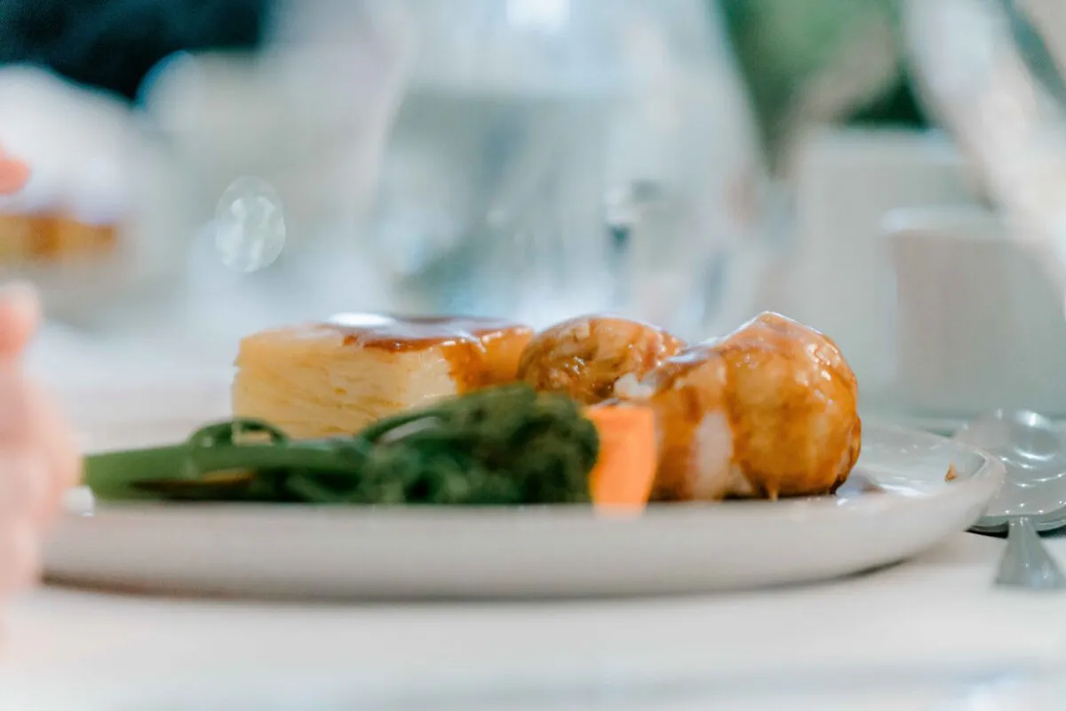 A beautifully plated wedding breakfast main course featuring roasted meat, layered potato, and green vegetables rests on a white plate under soft natural light.