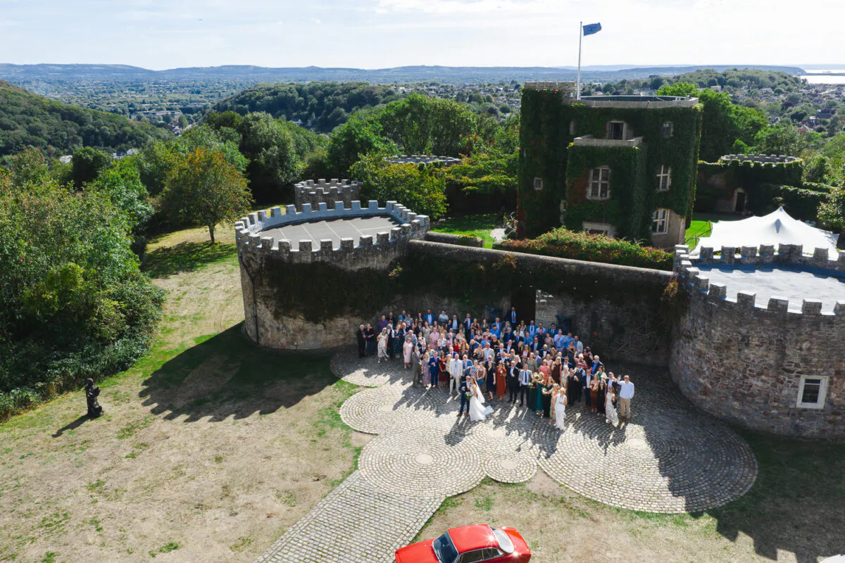 An aerial drone shot captures a large wedding party gathered in the cobbled courtyard of a historic stone castle surrounded by lush green countryside.