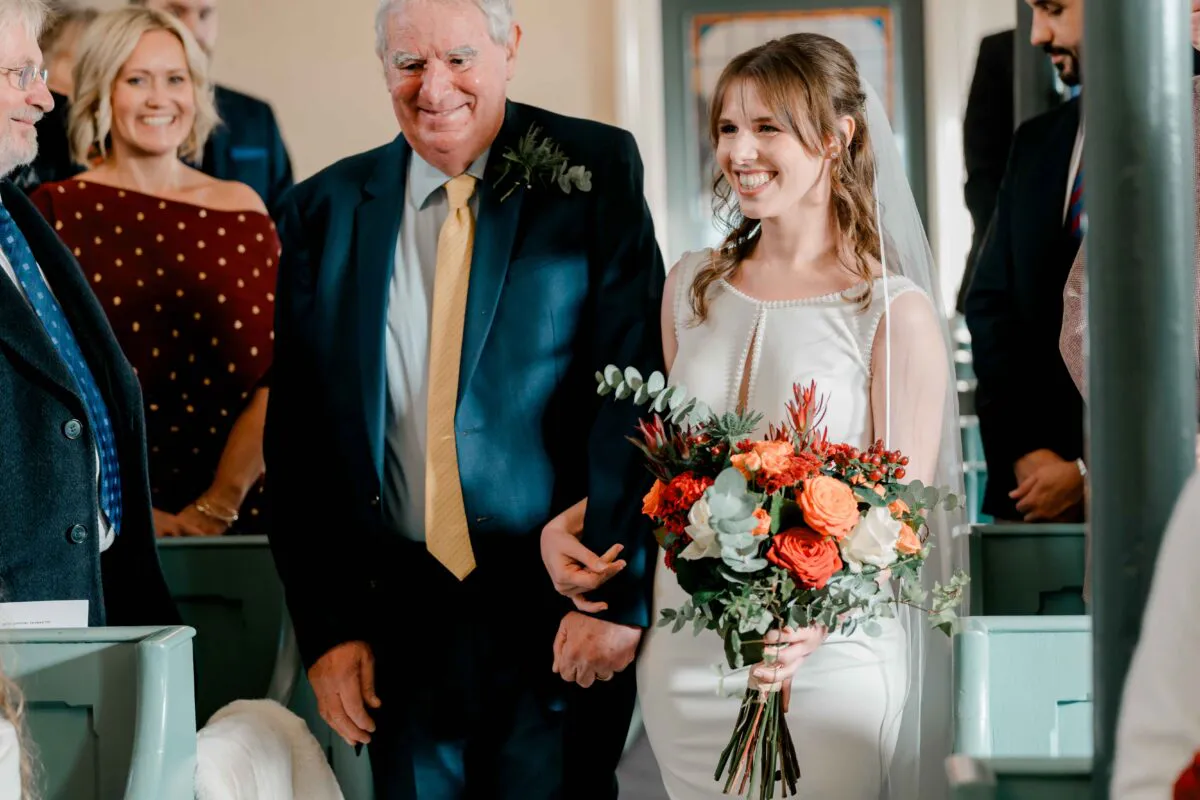 A smiling bride walks down the aisle arm in arm with her father, holding a vibrant autumn-toned bouquet filled with orange roses and eucalyptus.