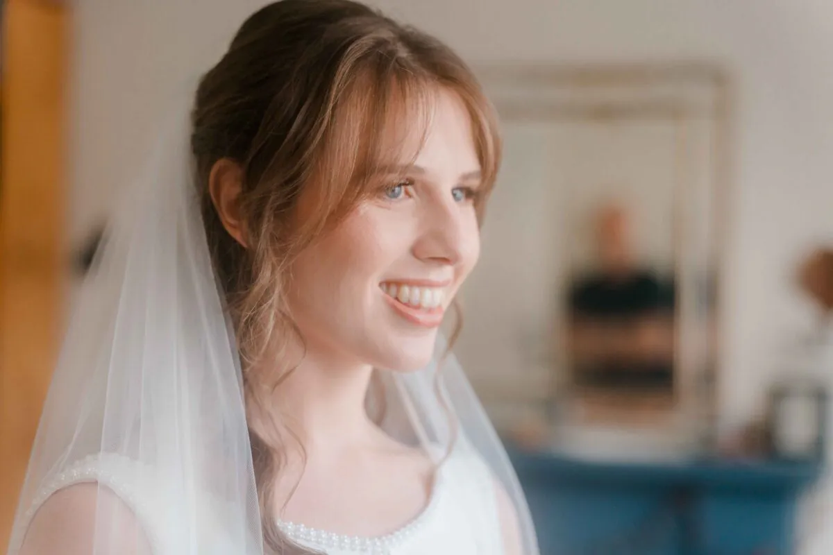 A smiling bride wearing a half-up hairstyle and veil looks towards a mirror during her wedding morning preparation.