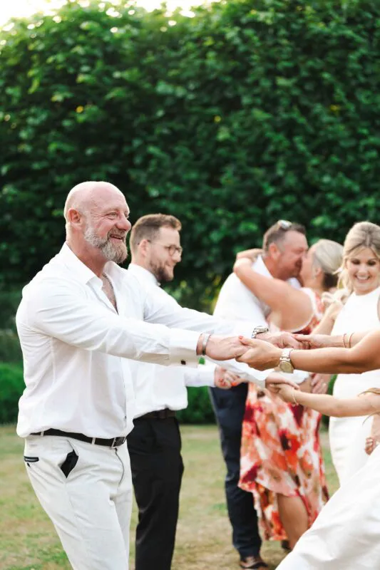 A joyful wedding guest laughing heartily while holding hands with others in a circle during an outdoor garden party celebration.