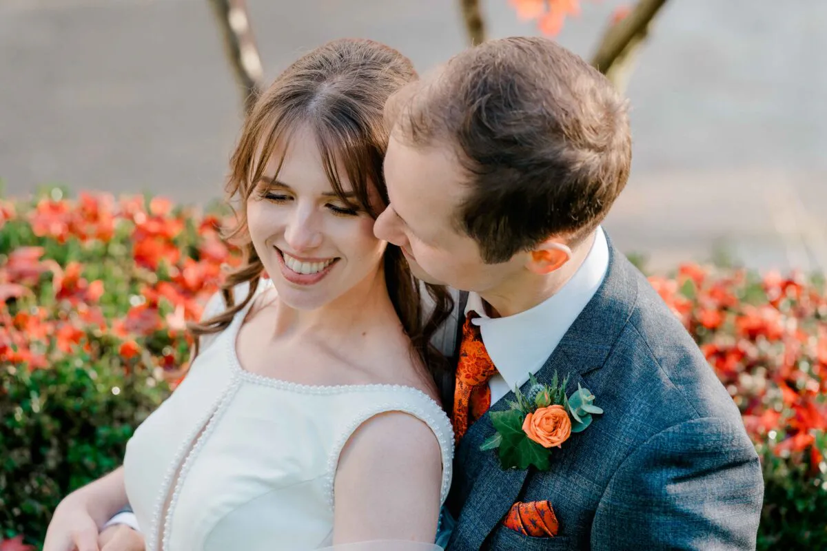 A smiling bride and groom share a tender moment outdoors with a vibrant red floral background framing them.