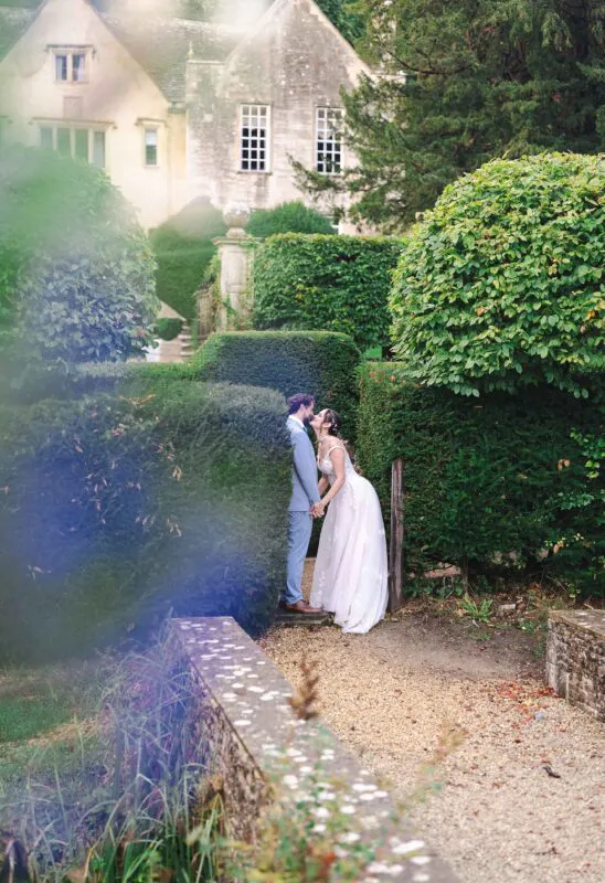 A newlywed couple shares a tender nose-to-nose moment amidst manicured topiary gardens outside a grand stone manor house on a sunny summer day.