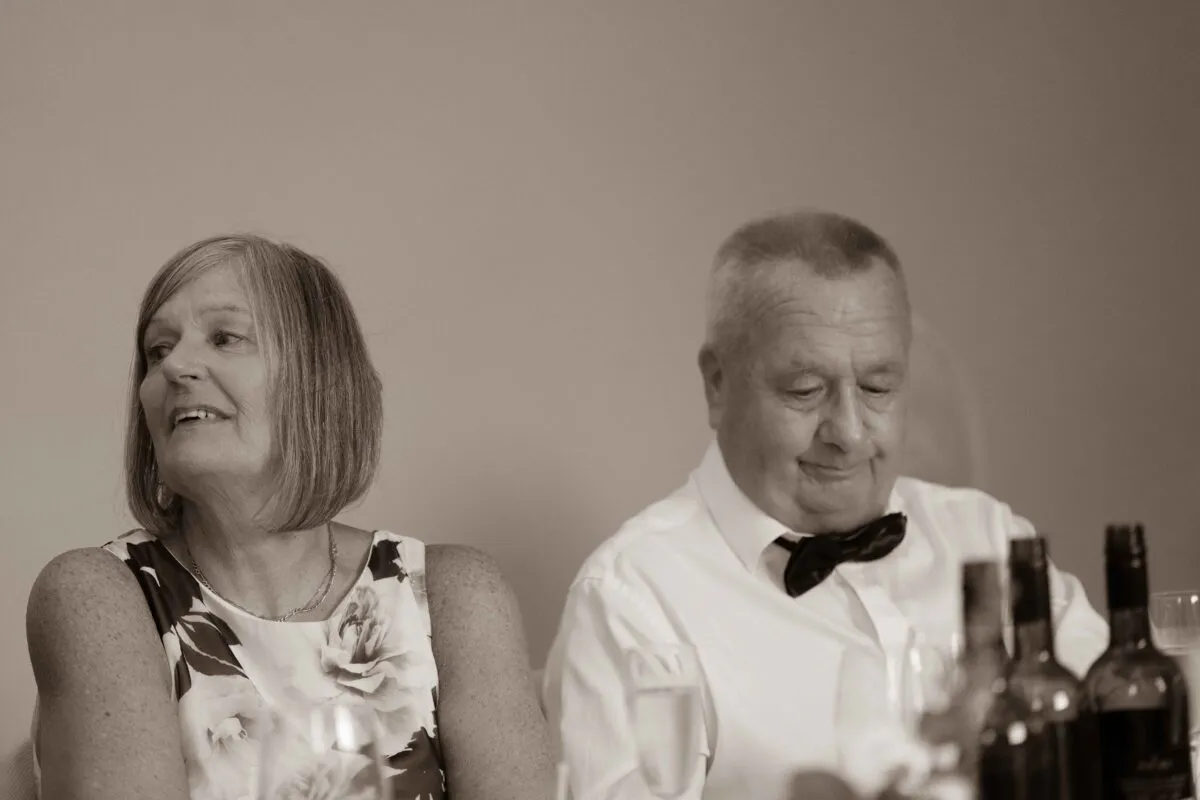 An older couple captured in a sepia-toned moment during the wedding reception speeches, with the woman smiling and looking away while the man looks down thoughtfully.