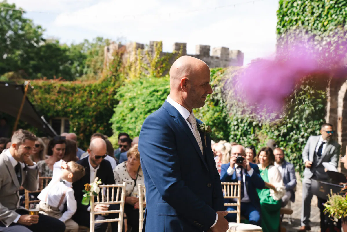 A groom in a smart navy suit stands smiling outdoors at an altar with seated guests and a castle wall backdrop.