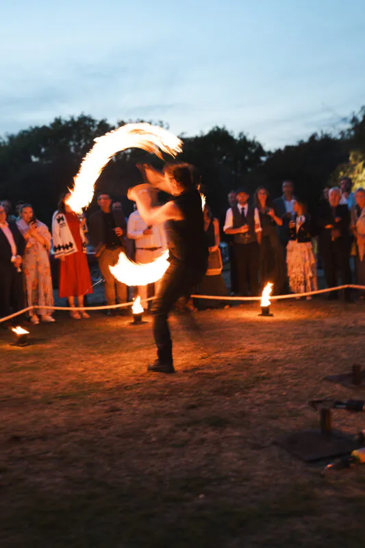 A fire performer creates sweeping arcs of flame with spinning props as wedding guests watch during an evening reception outdoors.