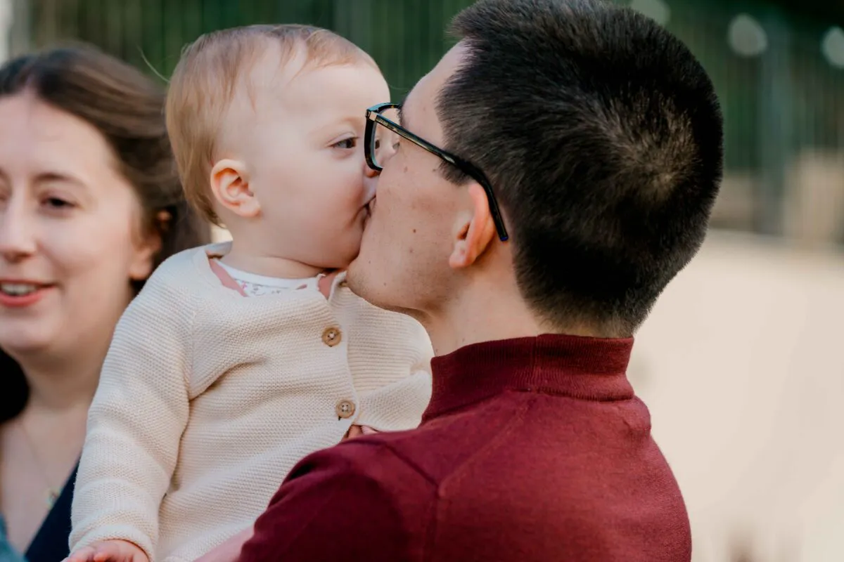 A close-up profile shot of a father lovingly kissing a baby while another woman smiles warmly nearby.