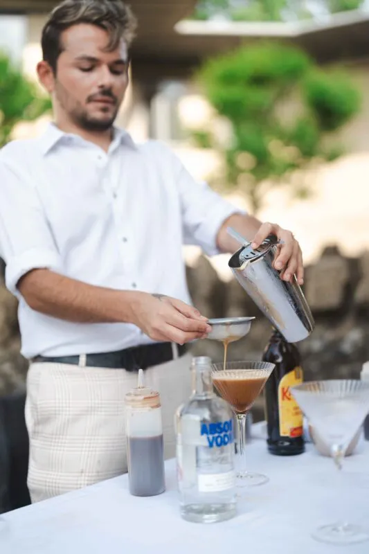 A professional mixologist carefully strains a freshly shaken espresso martini into a coupe glass at an outdoor wedding drinks reception.