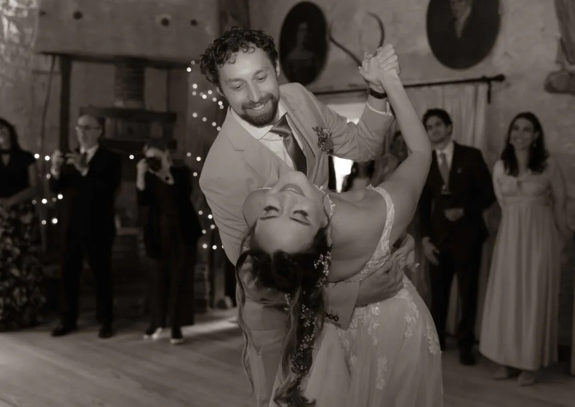 The newly married couple share a joyous dip during their first dance inside a rustic barn venue while guests watch on happily.