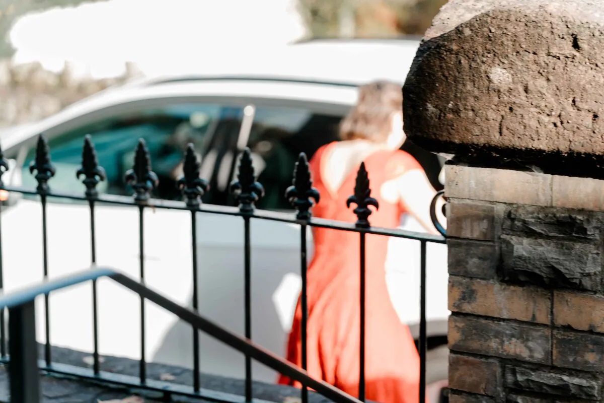 A guest in a vibrant red dress is seen from behind as she prepares to get into a car near a brick pillar and iron fence.
