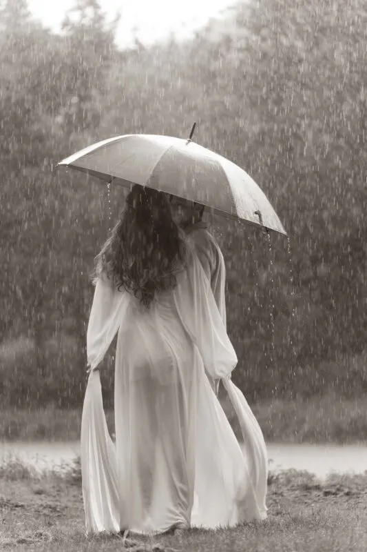 A newlywed couple stands closely under an umbrella amidst heavy rainfall in a sepia-toned outdoor portrait.