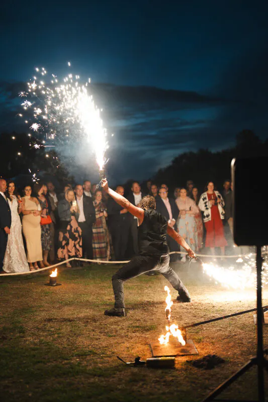 A fire performer entertains evening guests with a dynamic sparkler display under a twilight sky during a wedding celebration.