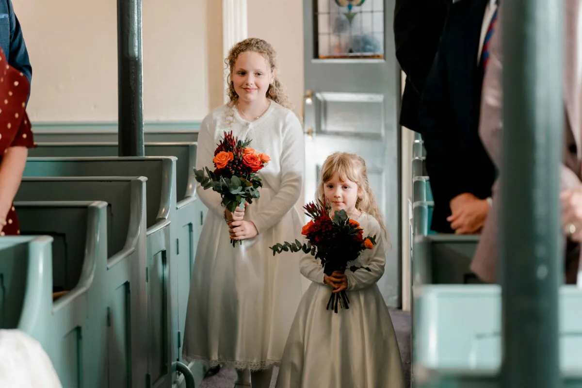 Two young flower girls in white dresses and cardigans carry small autumn-themed bouquets while walking down the aisle between green church pews.