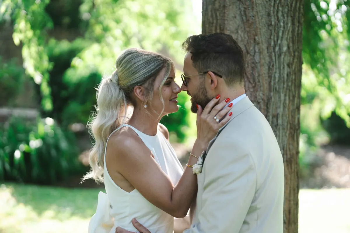 Newlyweds share an intimate nose-to-nose moment in a sun-dappled woodland, with the bride wearing a sleek high-neck gown and the groom in a classic beige suit.