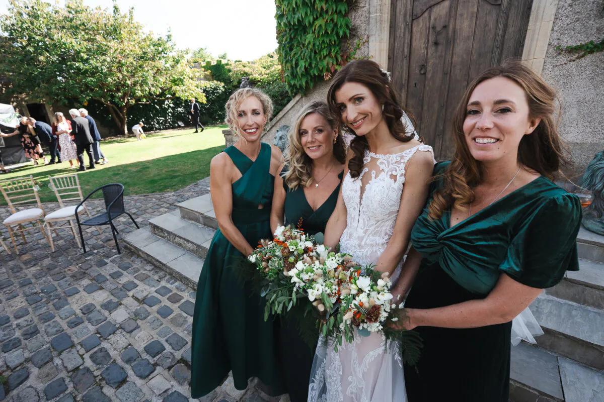 A smiling bride holding a hand-tied floral bouquet poses on a cobbled courtyard with three bridesmaids wearing elegant green dresses.