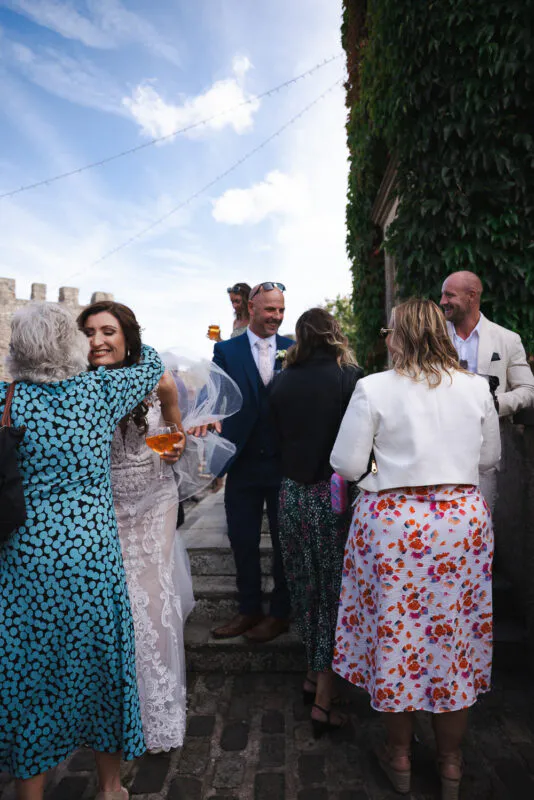 A bride in a lace gown embraces a guest during an outdoor drinks reception in a cobbled courtyard framed by an ivy-covered stone wall.