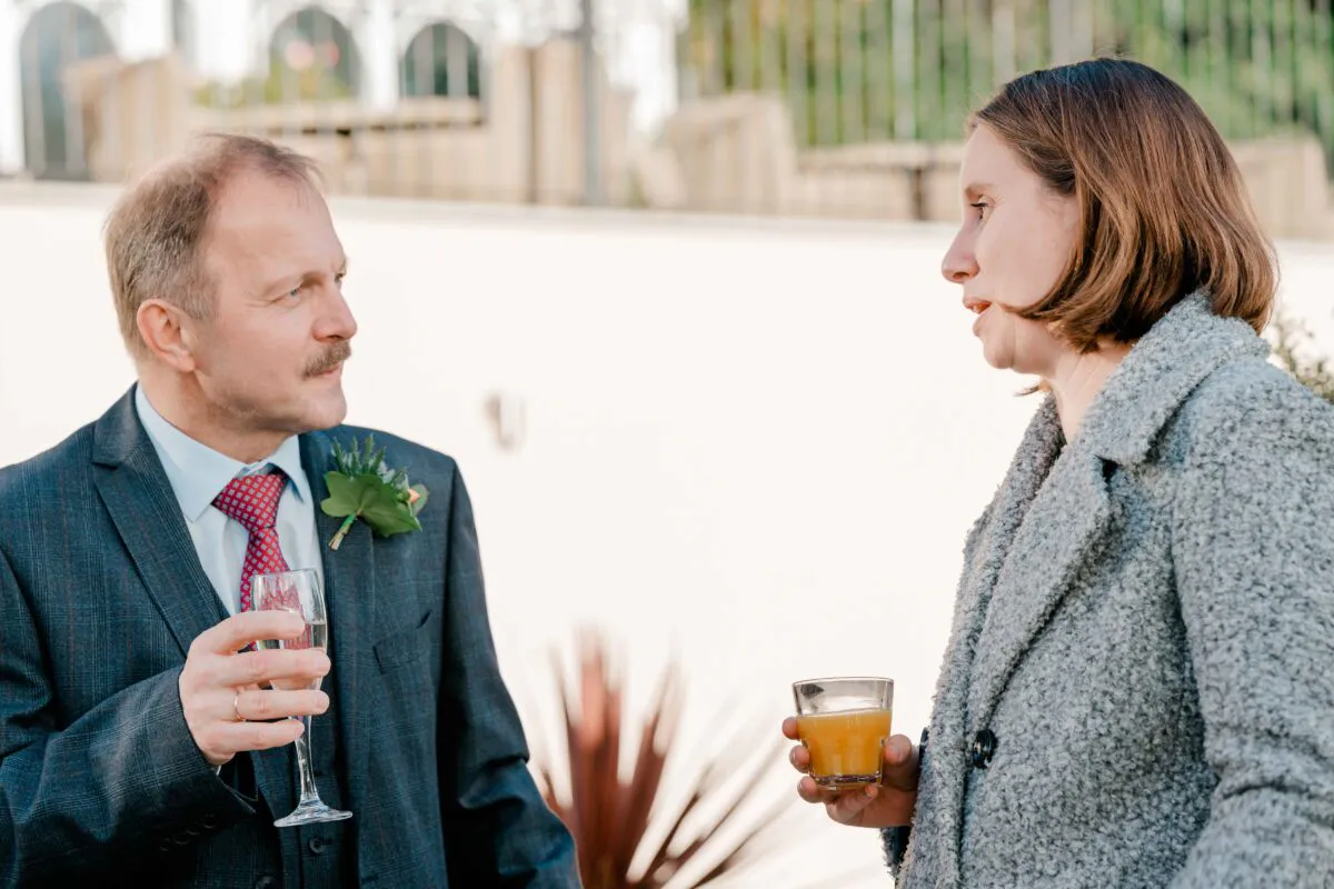 A mature wedding guest in a checked suit with a boutonniere chats with a woman holding a drink during an outdoor drinks reception.