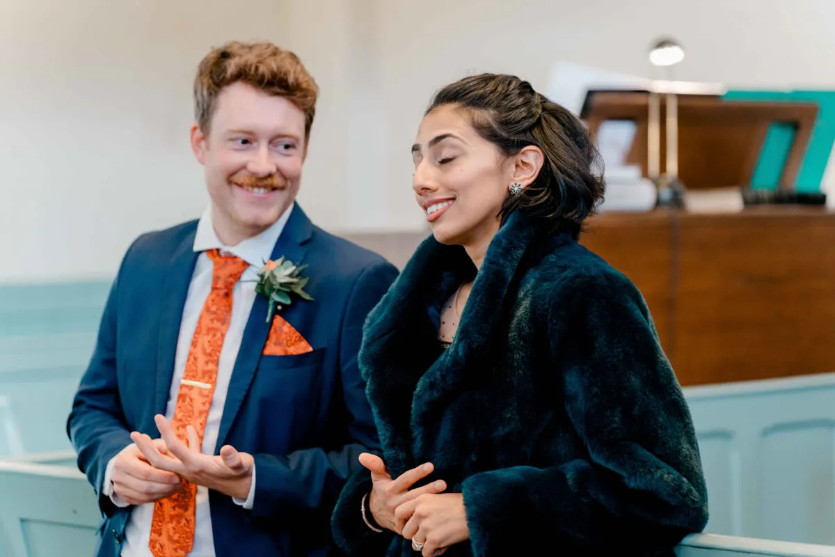 A smiling bride wearing a dark faux fur stole and a groom in a navy suit with an orange patterned tie stand together during an indoor ceremony.