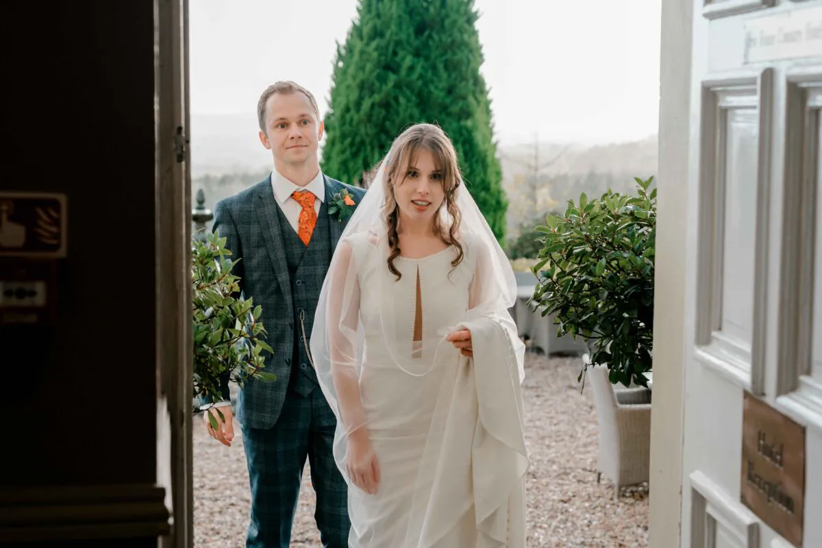 A newlywed couple pauses in a doorway, with the bride wearing a sleek dress and veil while the groom in a tweed suit stands behind her.