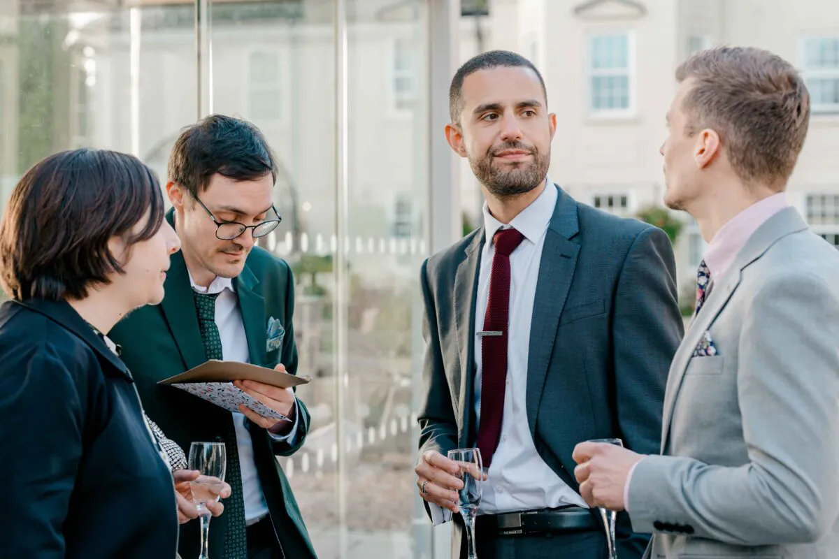 A group of wedding guests mingle and chat happily during the drinks reception, holding glasses of champagne while dressed in smart suits.