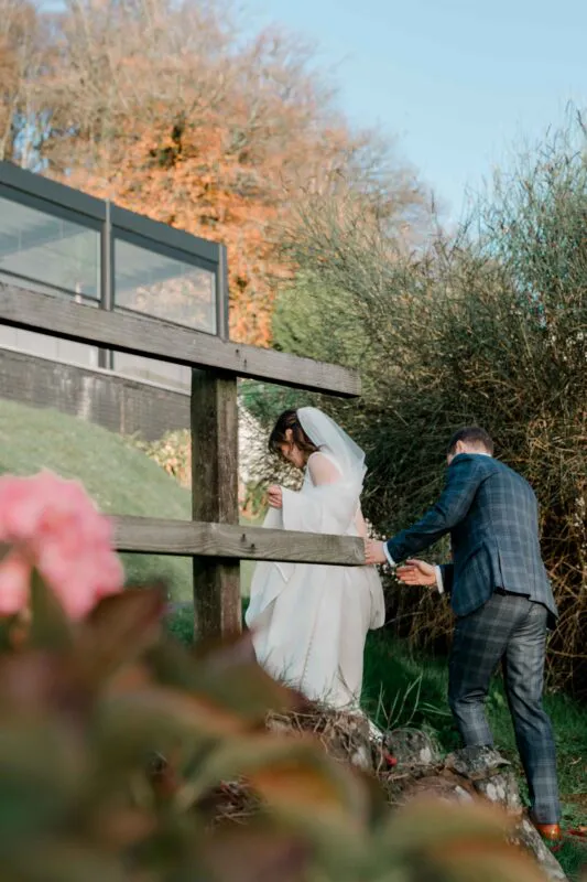 A bride and groom walk together outdoors near a rustic wooden fence in a natural, tree-lined setting during an autumn wedding.