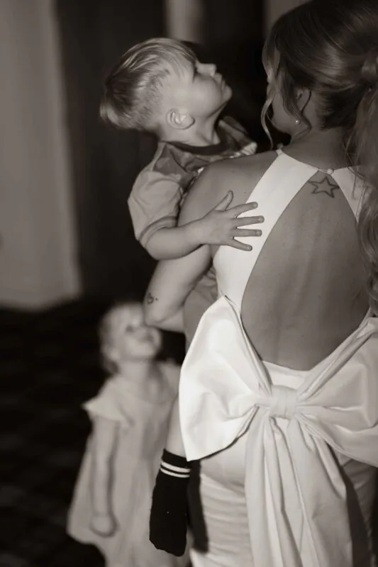 A bride in a backless gown with an oversized bow holds a young child in her arms during a tender moment.