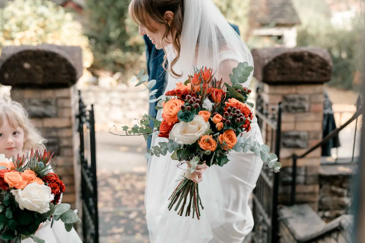 A joyful bride wearing a white gown and veil holds a vibrant autumn-toned bouquet while walking near stone pillars alongside a young flower girl.